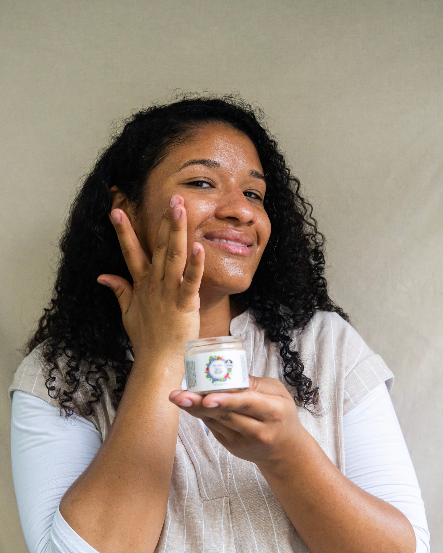A smiling woman holds a jar of the Sweet Dreams Glow Balm while applying it to her face.