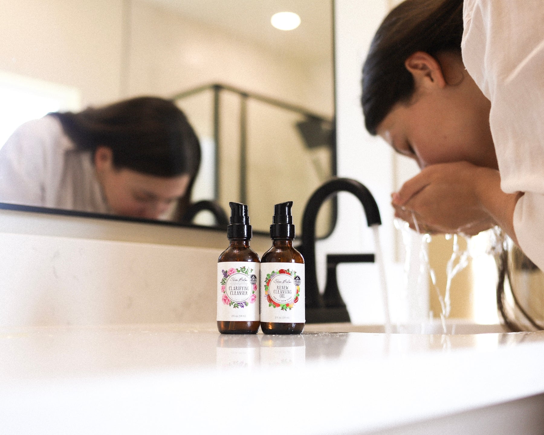 Facial Cleansing Duo resting on a bathroom sink while a woman washes her face in the background.