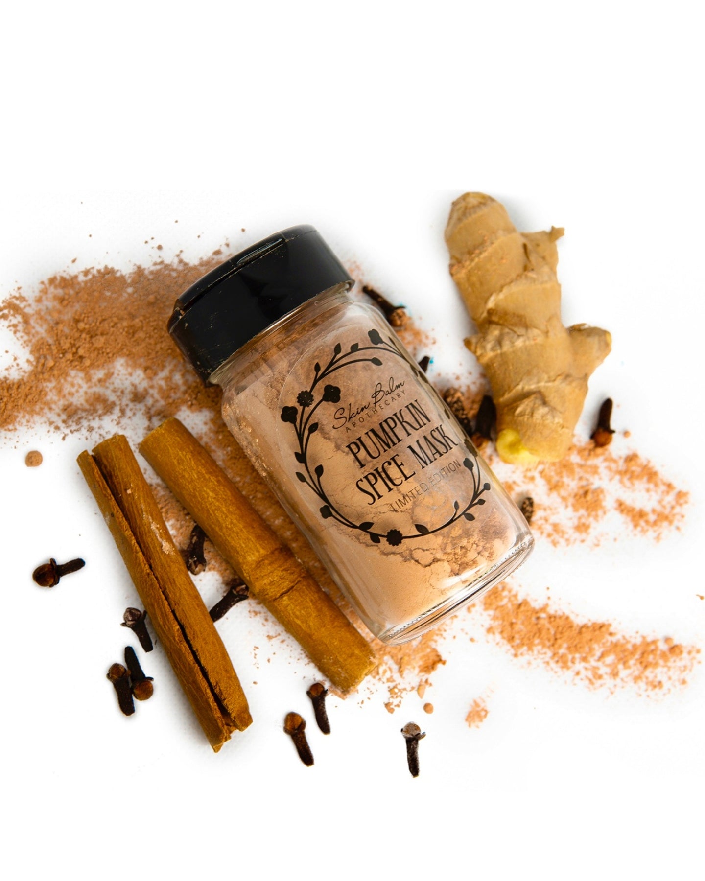 A spilled jar of Pumpkin Spice Mask surrounded by cinnamon sticks, ginger root, and clove buds against a white background.