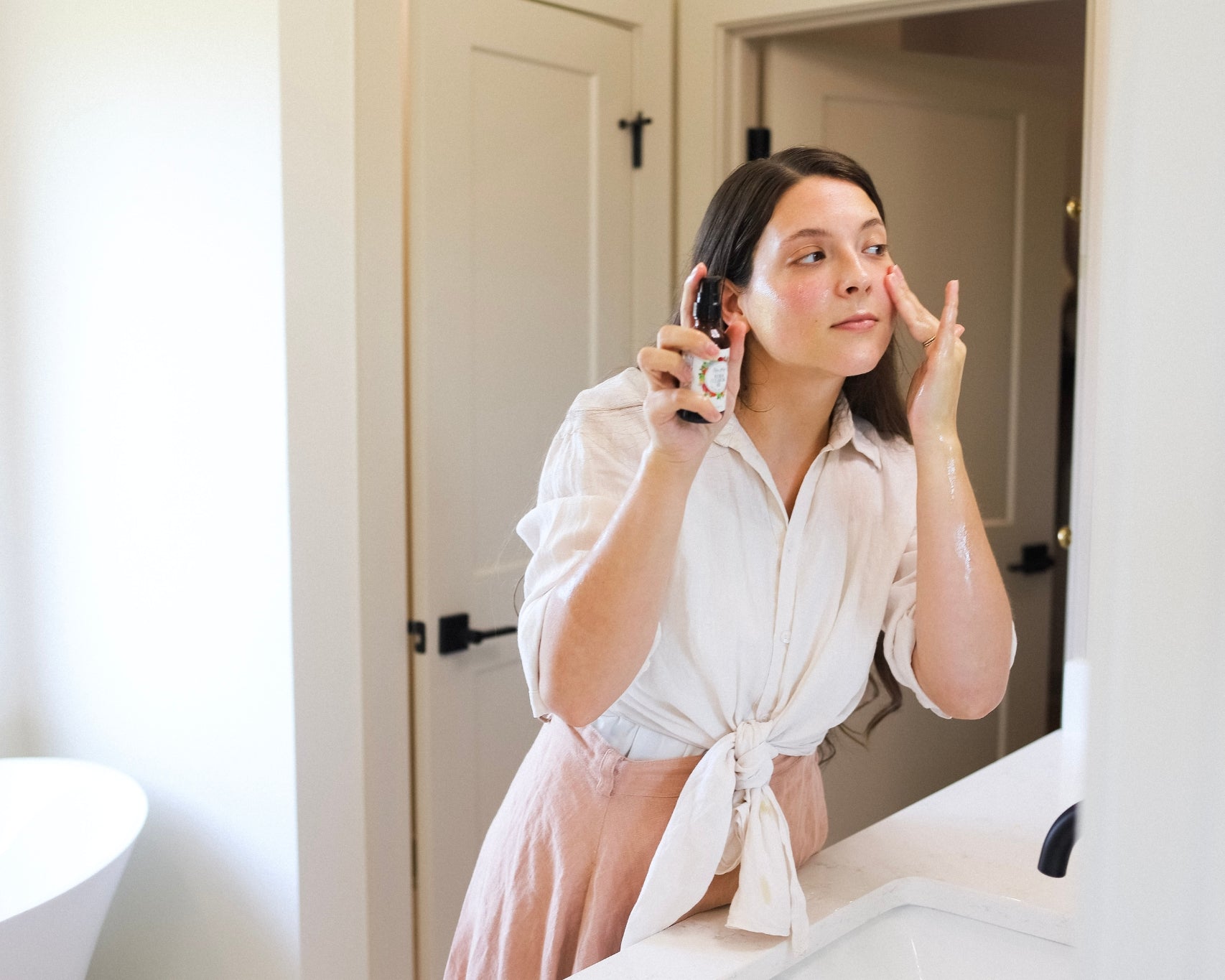 A woman holds up the Renew Cleansing Oil and rubs it onto her face while looking in the bathroom mirror.