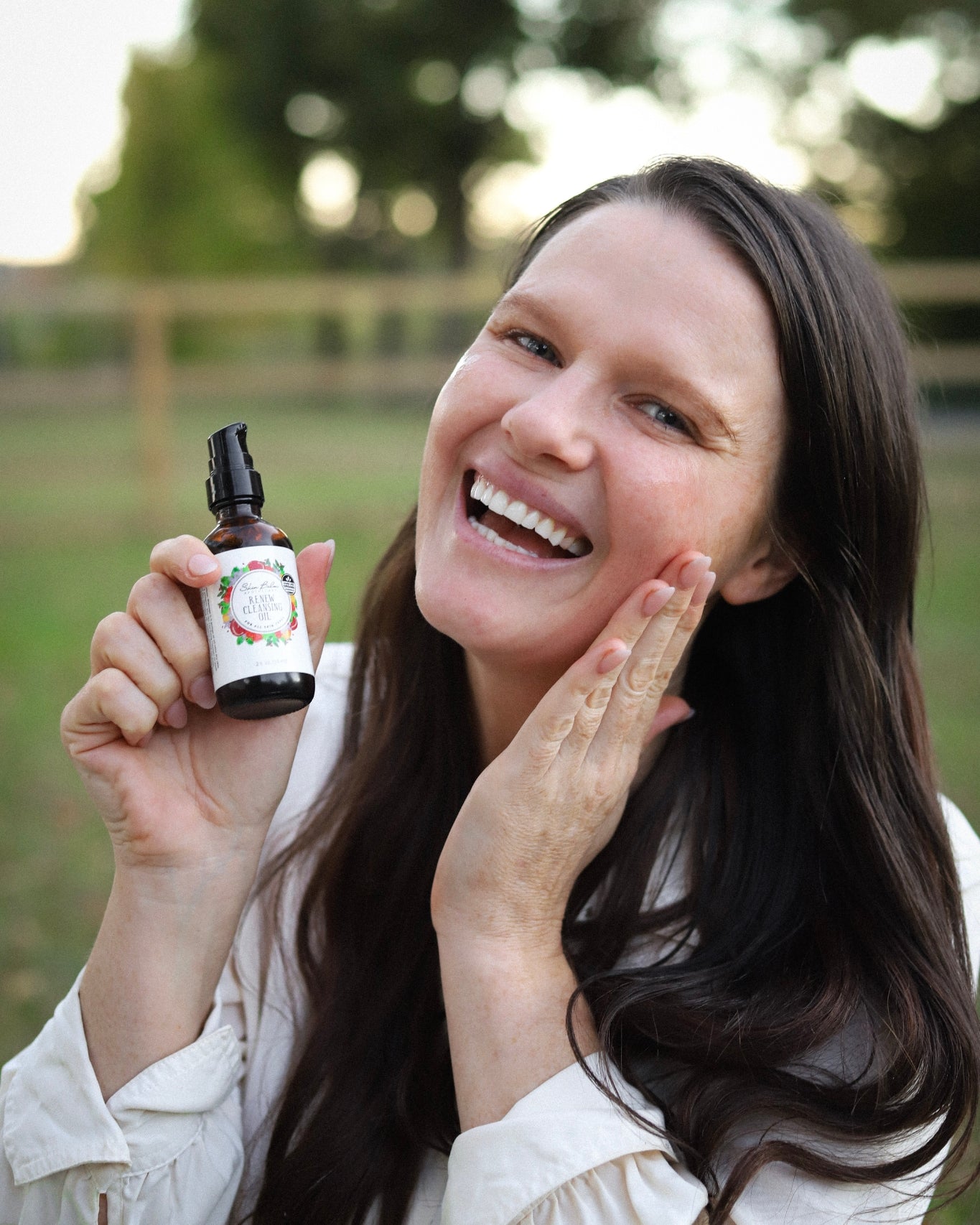 A smiling woman holds the Renew Cleansing Oil as she rubs it onto her face.